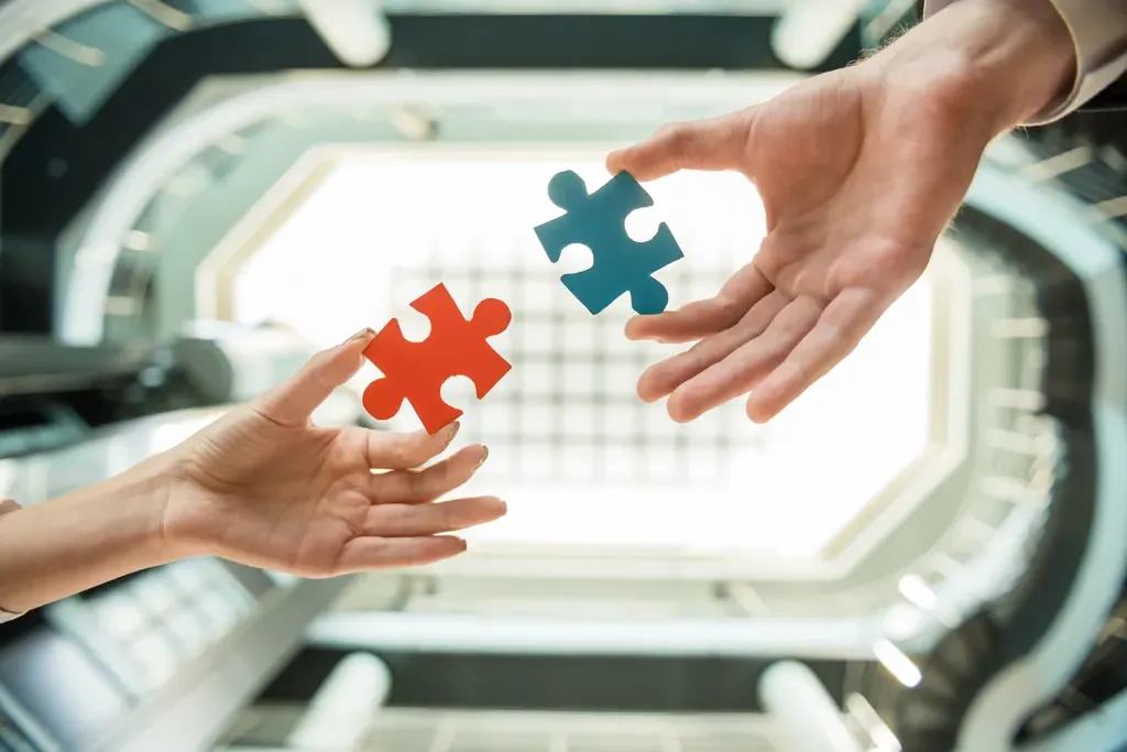 Deux mains tendant chacune un morceau de puzzle, un rouge et un bleu, prêtes à s'assembler, avec un fond flou de bâtiment moderne.