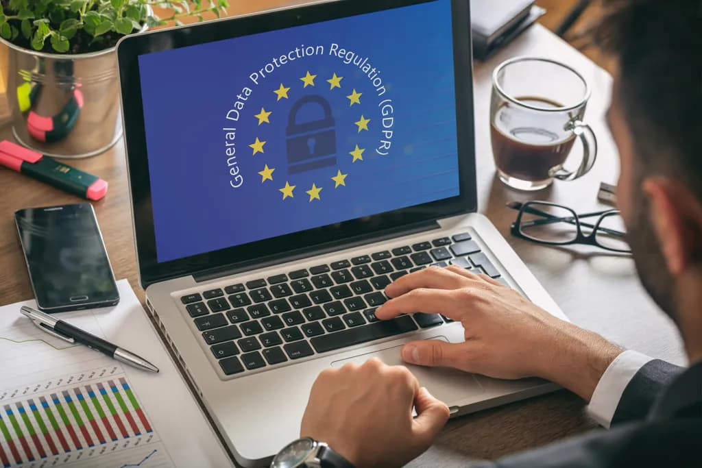 Une personne travaillant sur un ordinateur portable avec un écran affichant 'General Data Protection Regulation (GDPR)'. La personne utilise un clavier et semble être dans un environnement de bureau avec des documents, un téléphone, des lunettes, une tasse de café et des plantes autour.